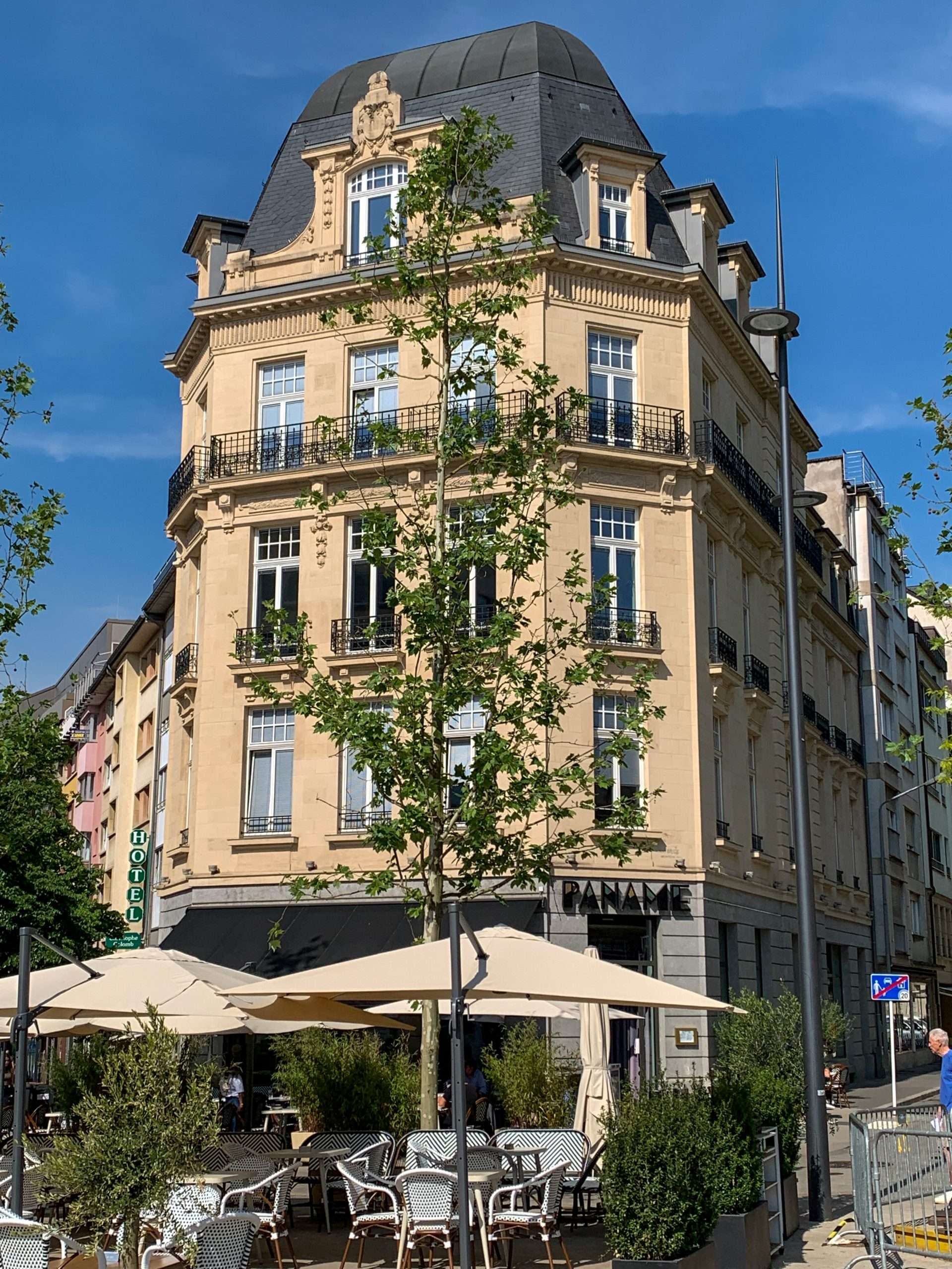 A café located on a street corner in Luxembourg city. A large green tree and café umbrellas are in the foreground whilst the building housing the café is in the background. The building has a definite Parisian style to it with multi stories and iron balconies. 