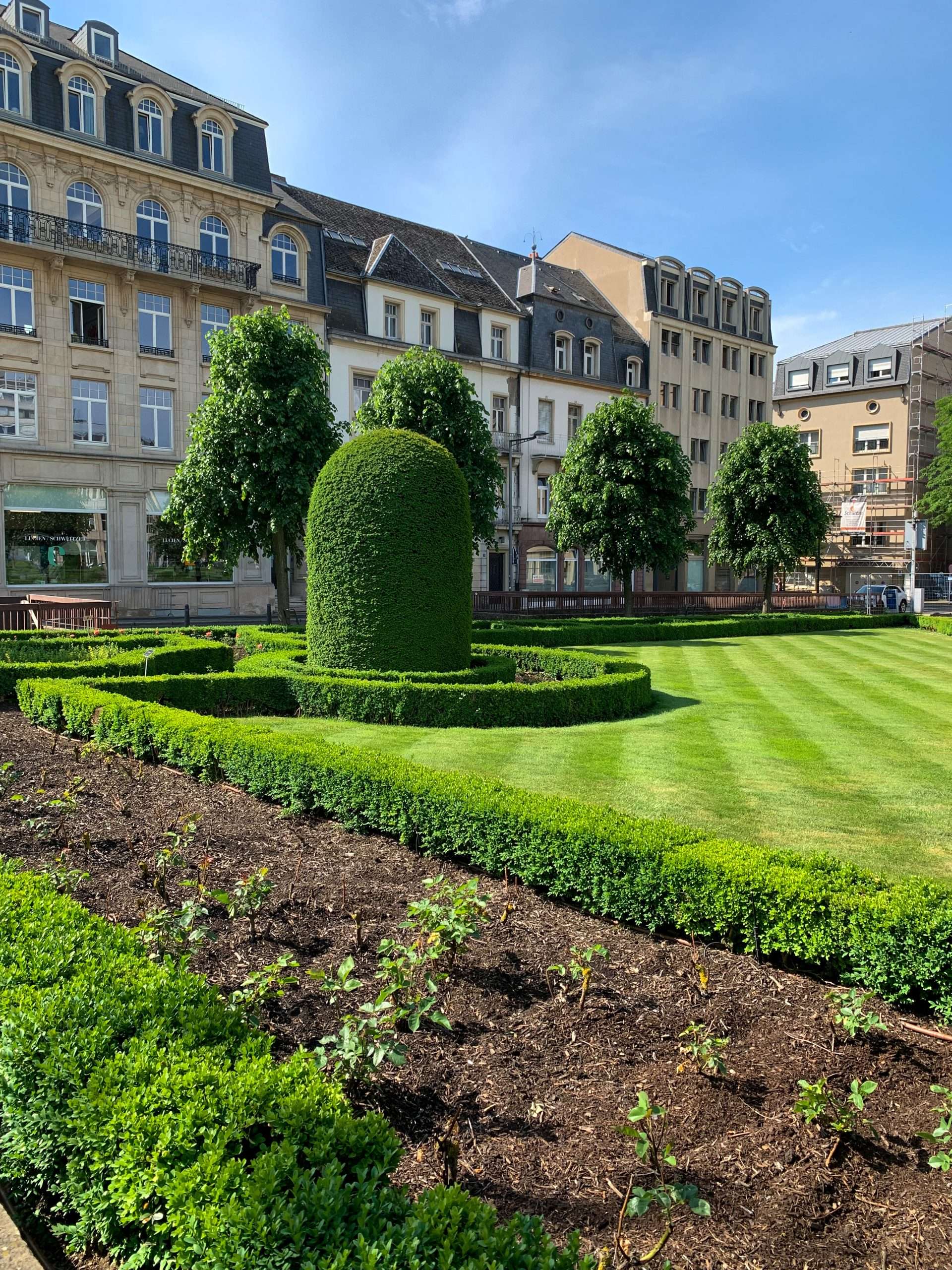 A view looking into the garden known as Place des Martyrs in Luxembourg city. Well manicured lawns and bushes are visible as are building on the streets surrounding the gardens. 