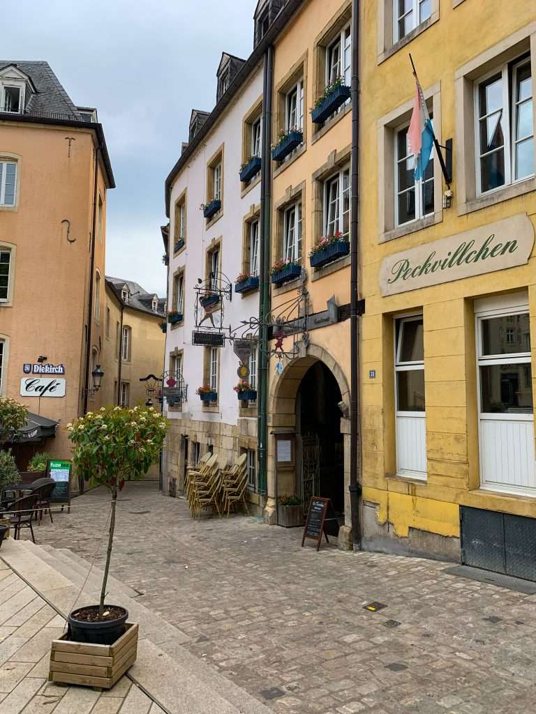 A street in Luxembourg's medieval old town can be seen together with some stacked chairs, a lemon tree in a pot and various flower boxes on surrounding buildings. The buildings are yellow, white and an orange colour.