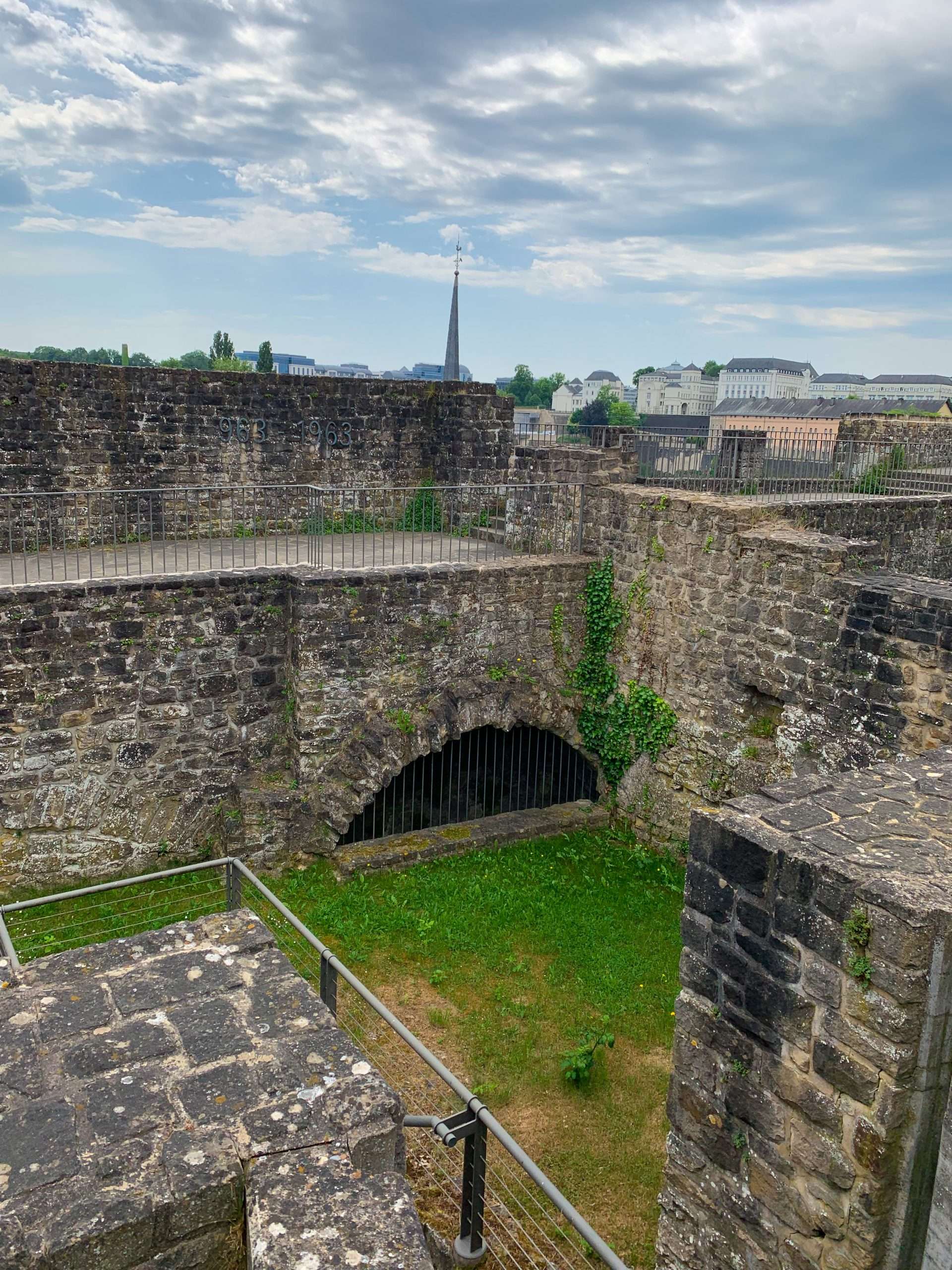 The Bock Promontory in Luxembourg with various walls and an arch visible in the image. Grass has grown amongst some of the walls and metal fencing has been installed for safety reasons. 