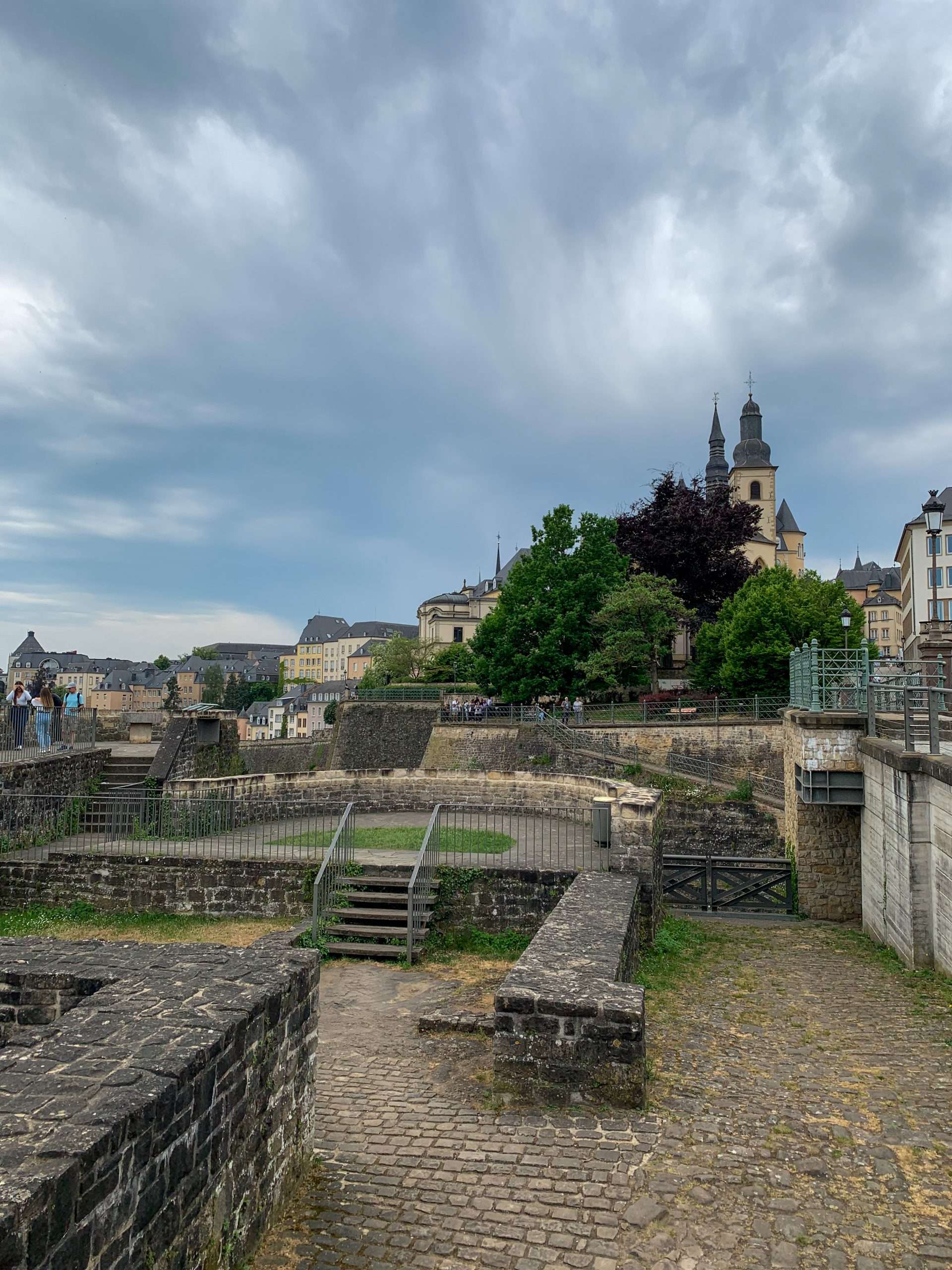 The Bock Promontory in Luxembourg with various stone structures visible in the image. Grass has grown amongst some of the walls and metal fencing has been installed for safety reasons. A view of the city of Luxembourg is visible in the background.