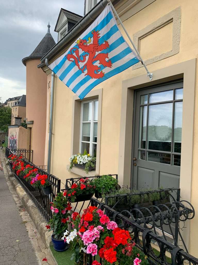 A view of the Luxembourg Flag hanging from a building in the old town of the city. Brightly coloured flowers of red, pink and white are in pots hanging from the iron fence in front of the building from which the flag hangs. 