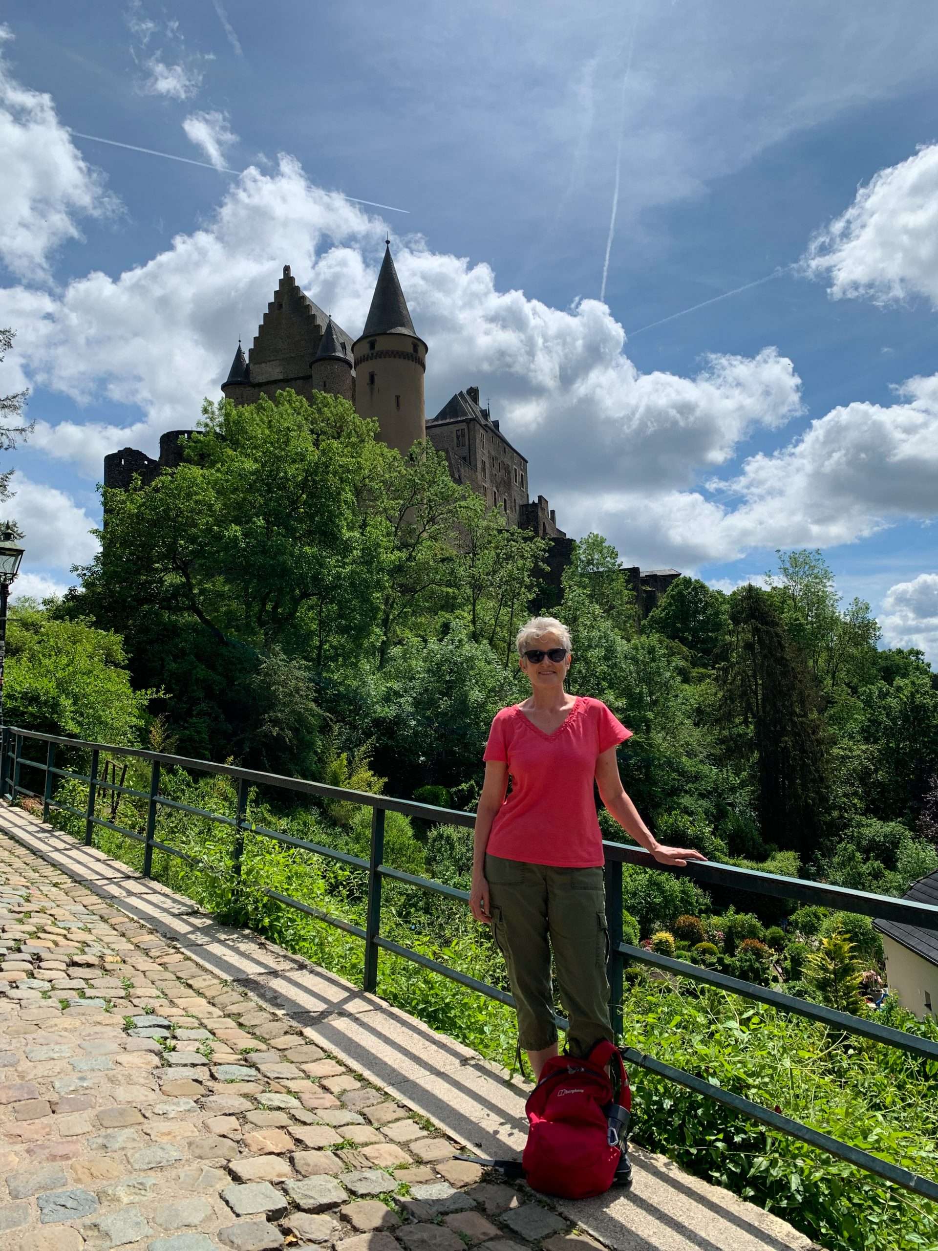 The author of this blog posts stands in front of Vianden Castle, Luxembourg. She is dressed in khaki colours trousers and an orange coloured short sleeved shirt. Her pink backpack sits in front of her at her feet.