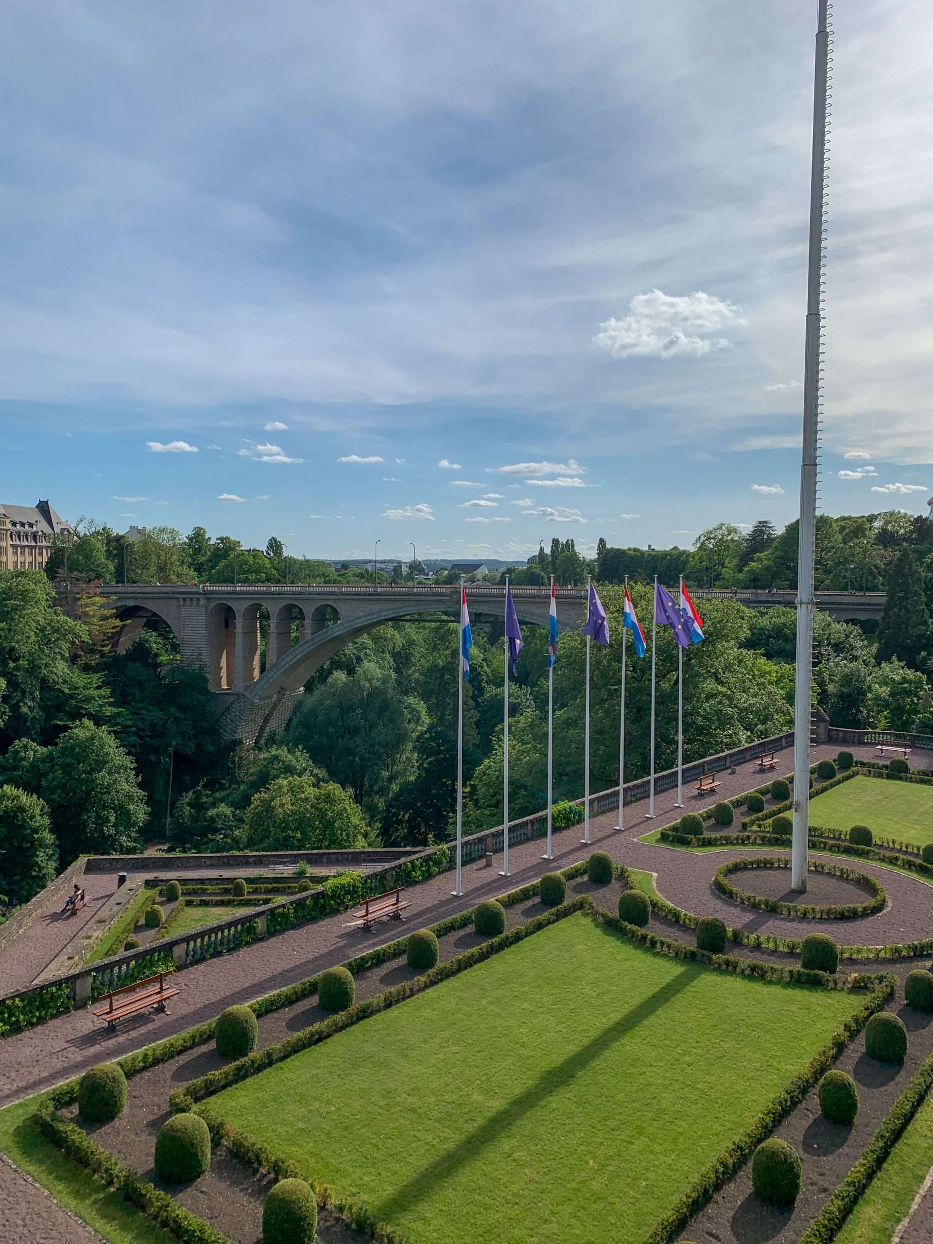 A view looking toward the Adolphe Bridge in Luxembourg. A manicured lawn, pathway, 7 flag poles with flags and part of a larger flagpole can be seen in the image. 