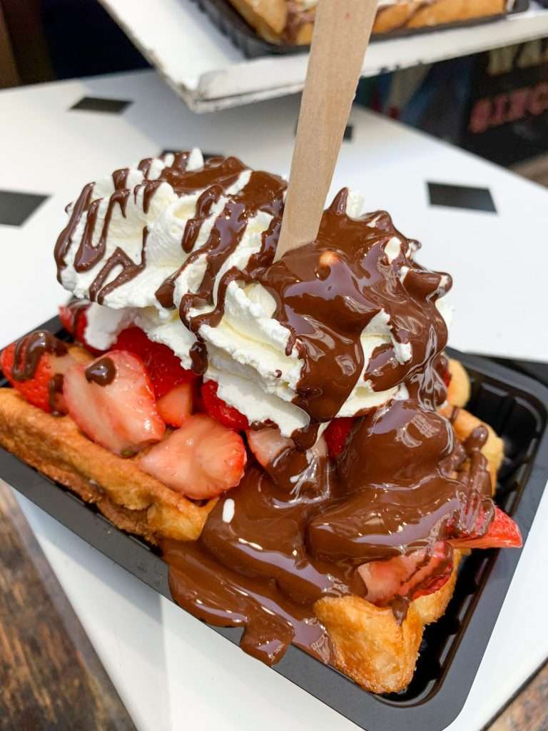 Image shows a Belgian waffle covered with cream, strawberries and melted chocolate 