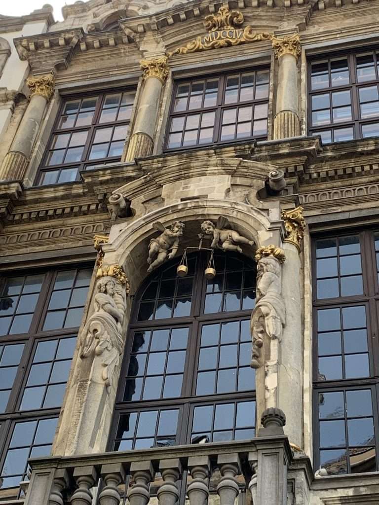 Image shows a building near the Grand Place in Brussels with a decorative façade including 2 cherubs holding a balanced scale.