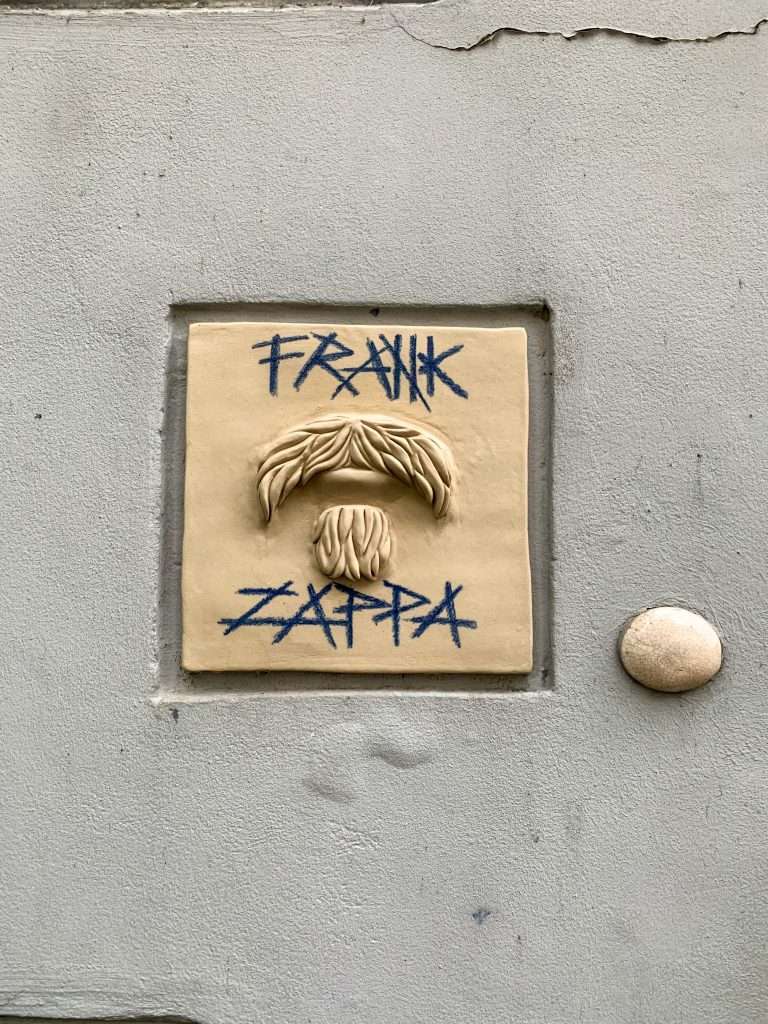 Image shows a small wall plaque with the words Frank Zappa written on them This can be seen along with other street art along Literatai Street in Vilnius