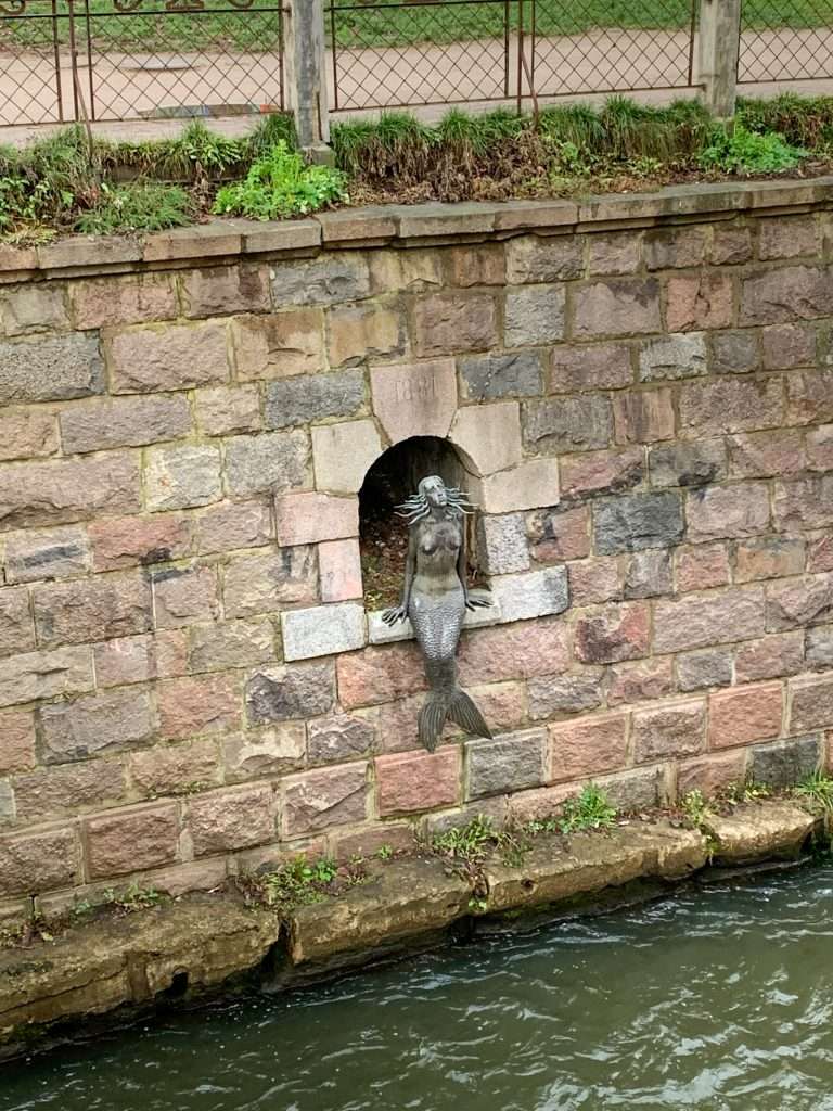 Image shows a statue called the Mermaid of Užupis located within the walls running alongside the Vilnia River which flows through the Republic of Užupis in Vilnius