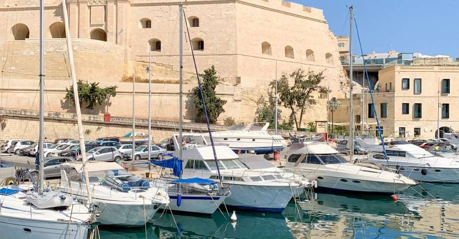 A view of Malta under a bright blue sky as seen from a boat in the harbour. Several moored boats are visible as are the golden coloured stone buildings around the harbour.