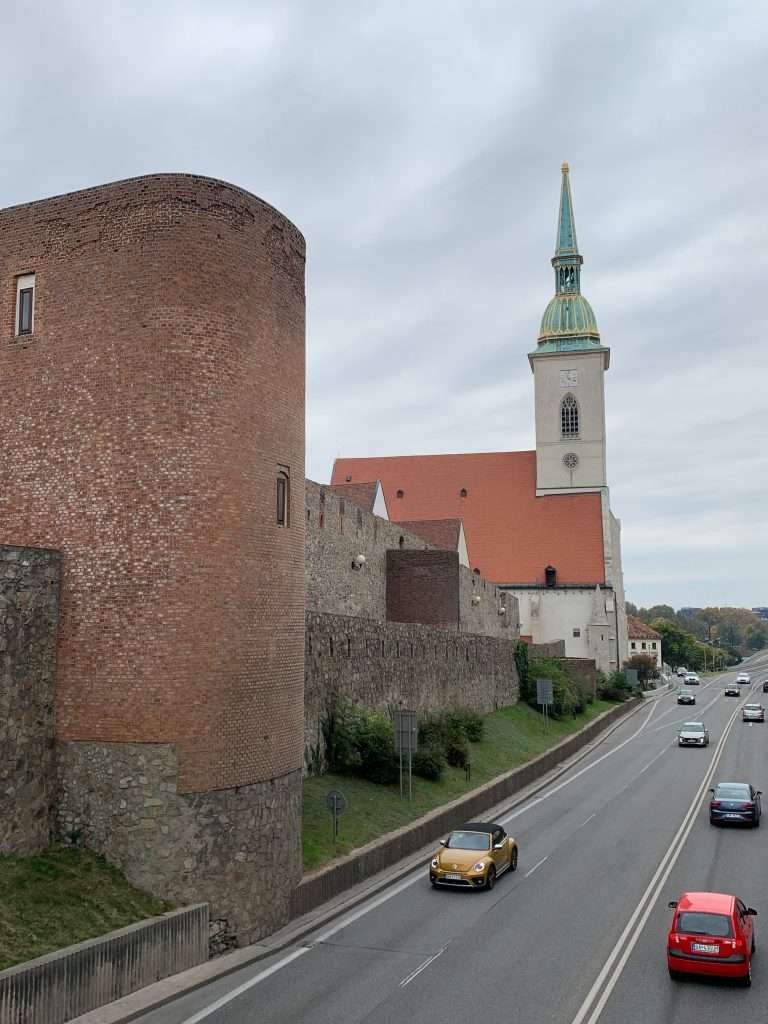 A view of the old town wall in Bratislava with the spire of St Martin's visible. Cars are seen travelling along the road which runs adjacent to the old town.