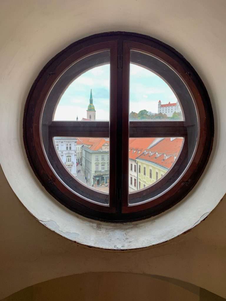 Image shows a view of the old buildings with Bratislava Castle in the distance. The image was taken from a window up in the Tower overlooking the Main Square in central Bratislava