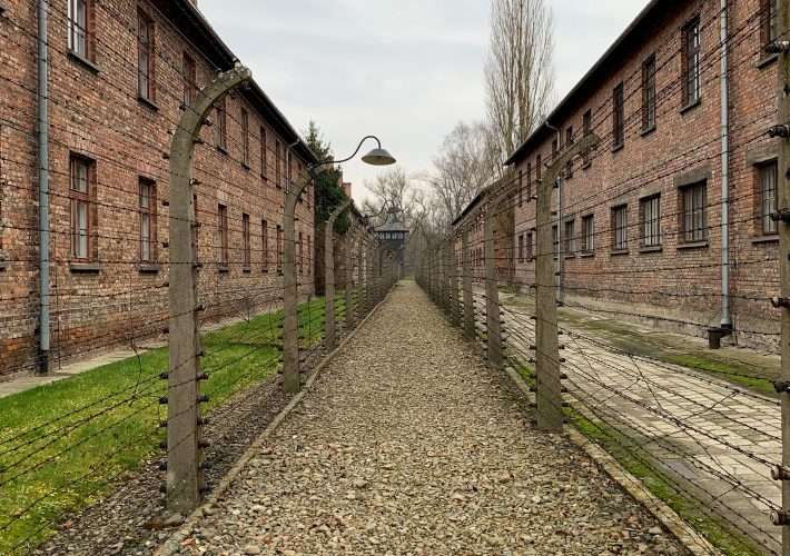 A single gravel path bordered with barbed wire fencing and long dormitory buildings with windows and forming part of the Auschwitz concentration camp near Krakow in Poland.