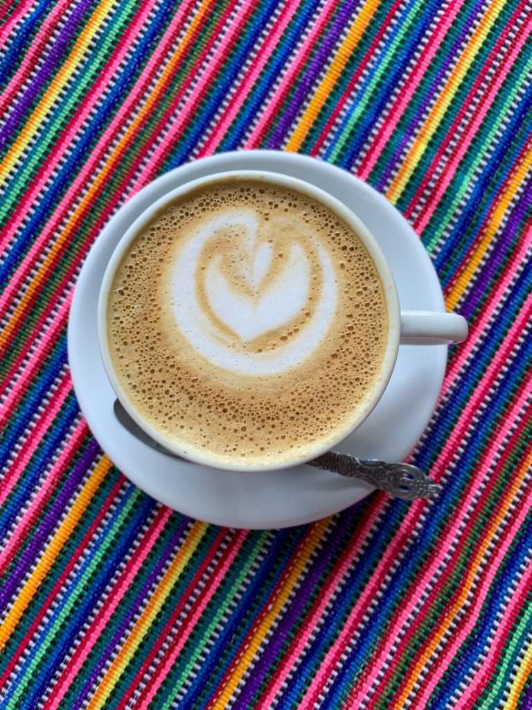 Image shows a cup of coffee against a brightly multi coloured striped table cover. Stopping for coffee is a must do in Antigua Guatemala!