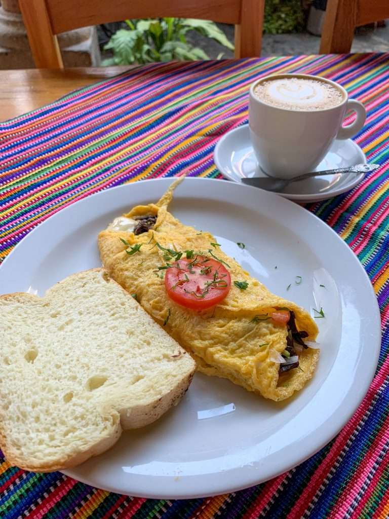 Image shows an omelette with bread and a cup of coffee against a brightly multi coloured striped table cover. Enjoying the local coffee and food is a must do in Antigua Guatemala!