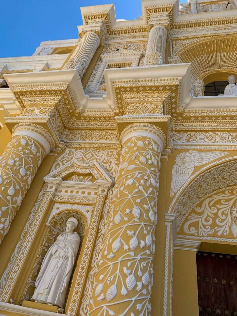 Image shows a close up view of the decorative white artwork on the outside of La Merced Church in Antigua Guatemala. The church is yellow in colour making the white artwork, which has the appearance of icing, stand out. This is a must visit site when spending a day in Antigua