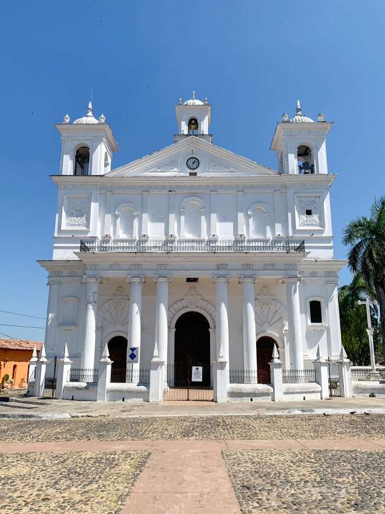 The white coloured Santa Lucia church sits beneath a bright blue Suchitoto sky. The cobbled area in front of the church is visible in the image.
