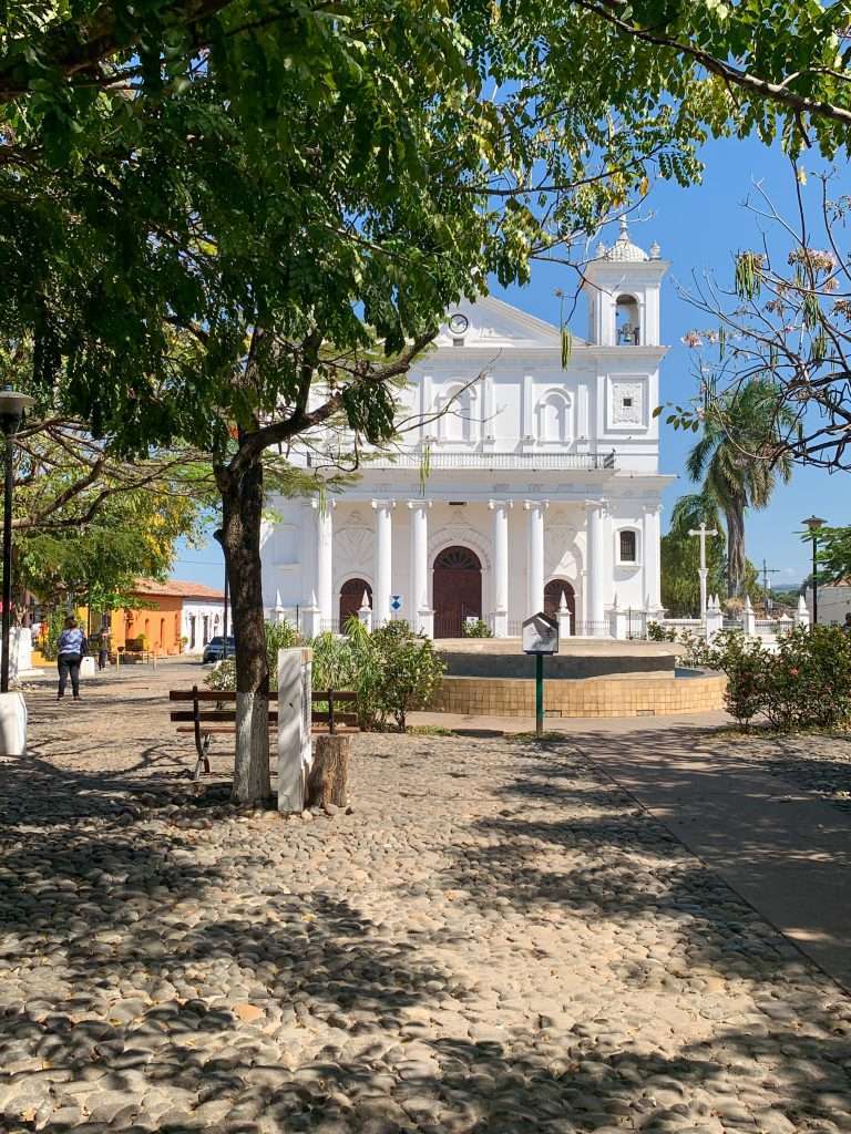 The white coloured Santa Lucia church sits beneath a bright blue Suchitoto sky with a fountain and trees visible in the foreground. The cobbled area in front of the church is visible in the image.