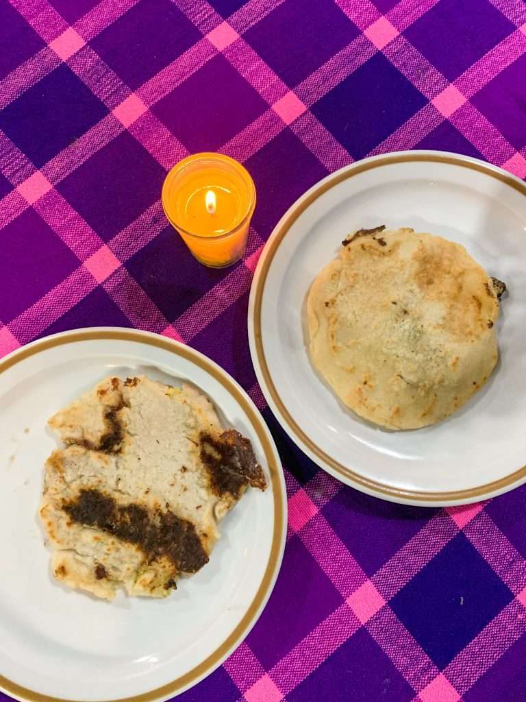 2 cooked pupusas each sit on a white plate with a purple and pink checked tablecloth beneath them. A small tea light candle burns next to the plates.