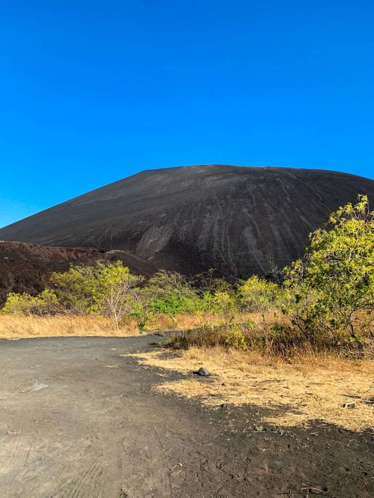 Cerro Negro, an active volcano in Nicaragua where volcano boarding takes place sits beneath a blue cloudless sky. Green but sparse vegetation is visible in the foreground with the black gravel of the volcano stark against the blue sky.