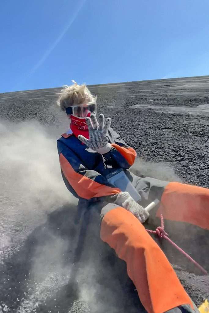 A close up view of the author of this blog post waving to the camera as she boards down the Cerro Negro active volcano in Nicaragua.