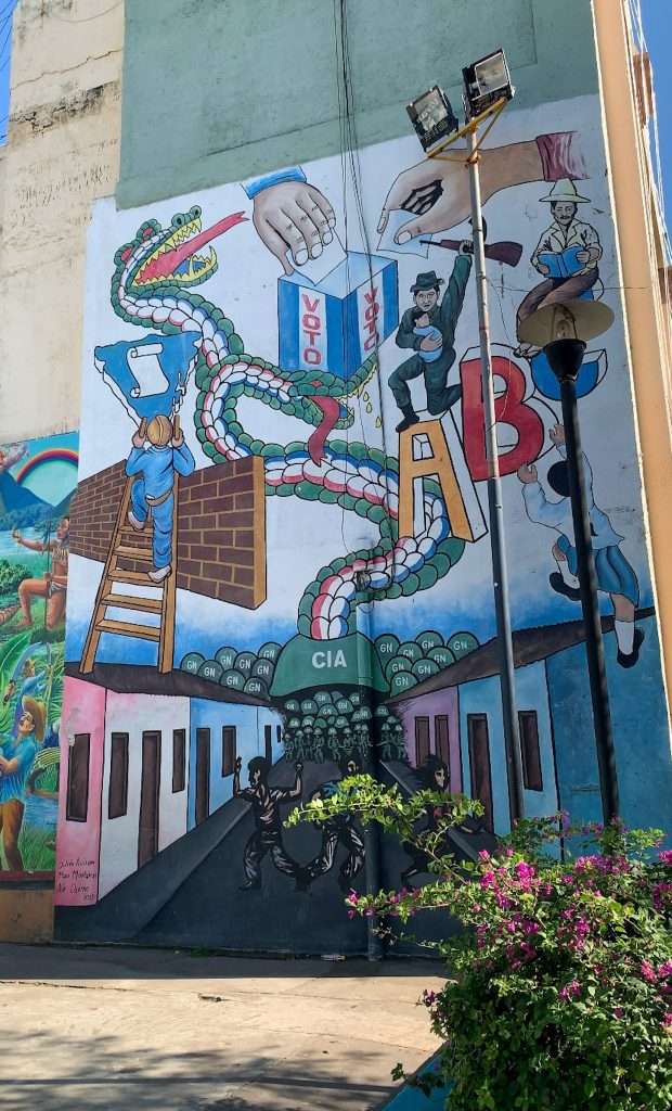 A multi coloured street mural in Leon Nicaragua depicting political themes including a voting box, dragon, the letters CIA and various people.
