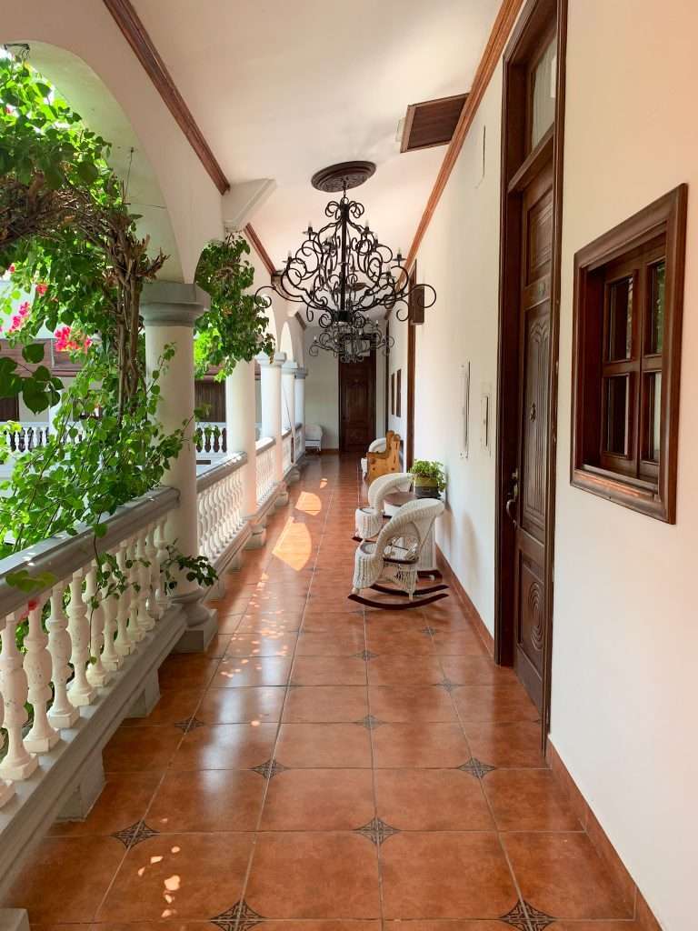 Aa view of the outside verandahs at the Hotel Real la Merced in Granada Nicaragua. Greenery frames the verandah in part and white rocking chairs can be seen outside individual hotel rooms.