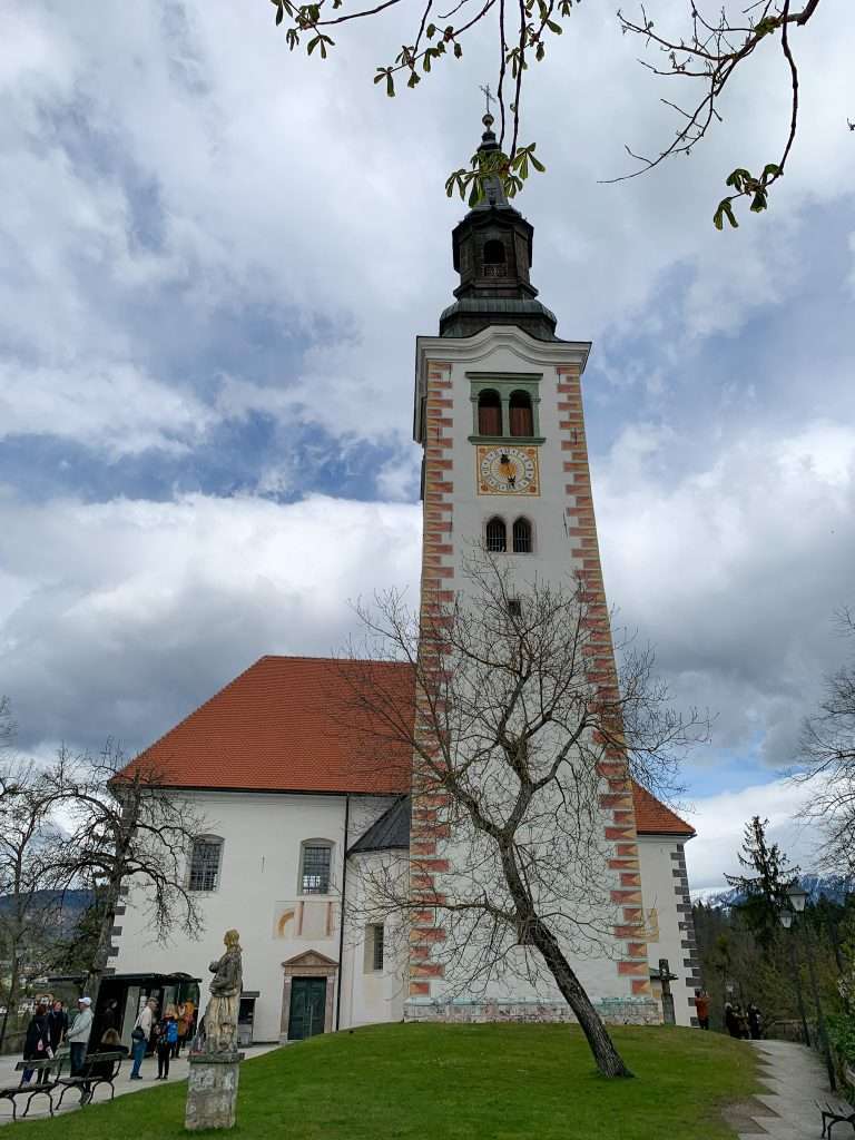 Thee Church on Bled Island stands on Bled Island. It is mainly white in color with a reddish coloured tiled roof and some reddish bricks around the corners of the tower.