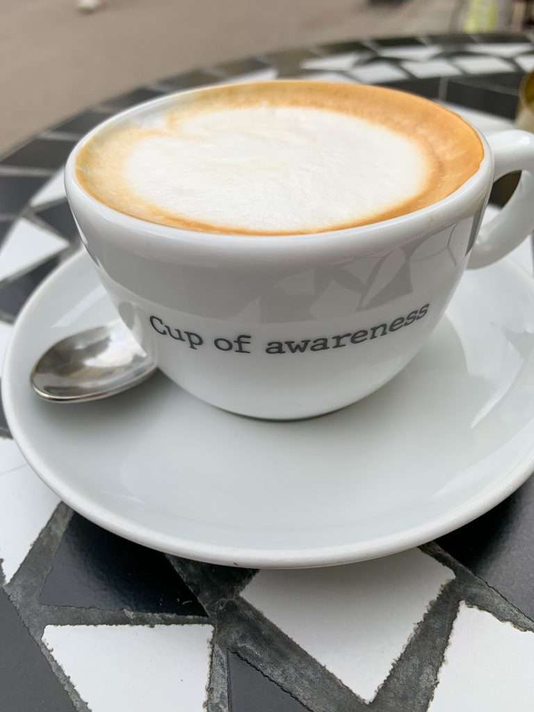 Coffee in one of Ljubljana's many lovely cafes and somewhere to go during a day in Ljubljana. This cup of coffee is in a white china cup with saucer underneath and the words "cup of awareness" are written in black on the cup.
