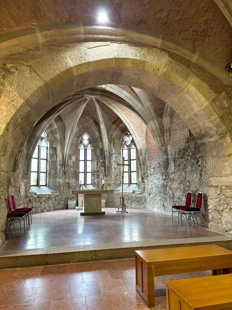 Photo of the inside of the Budapest history museum. The arched ceiling of a former chapel is visible as are some simple wooden benches. 