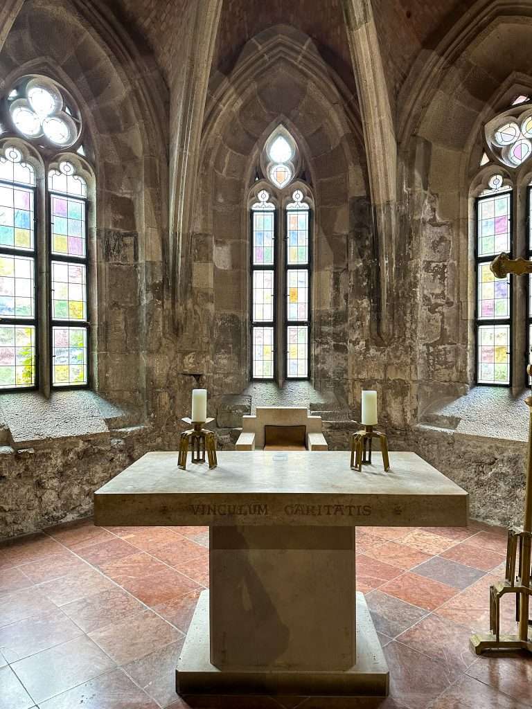 A close view of a stone table with 2 candles on top of it. The table is located beneath a medieval archway inside a chapel. 