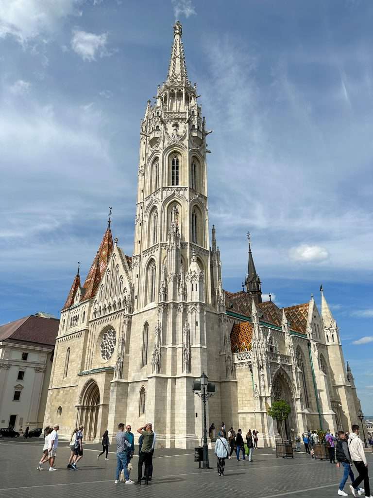 An external view of Mátyás Church in Budapest. Numerous people can be seen walking around the outside of the church.  