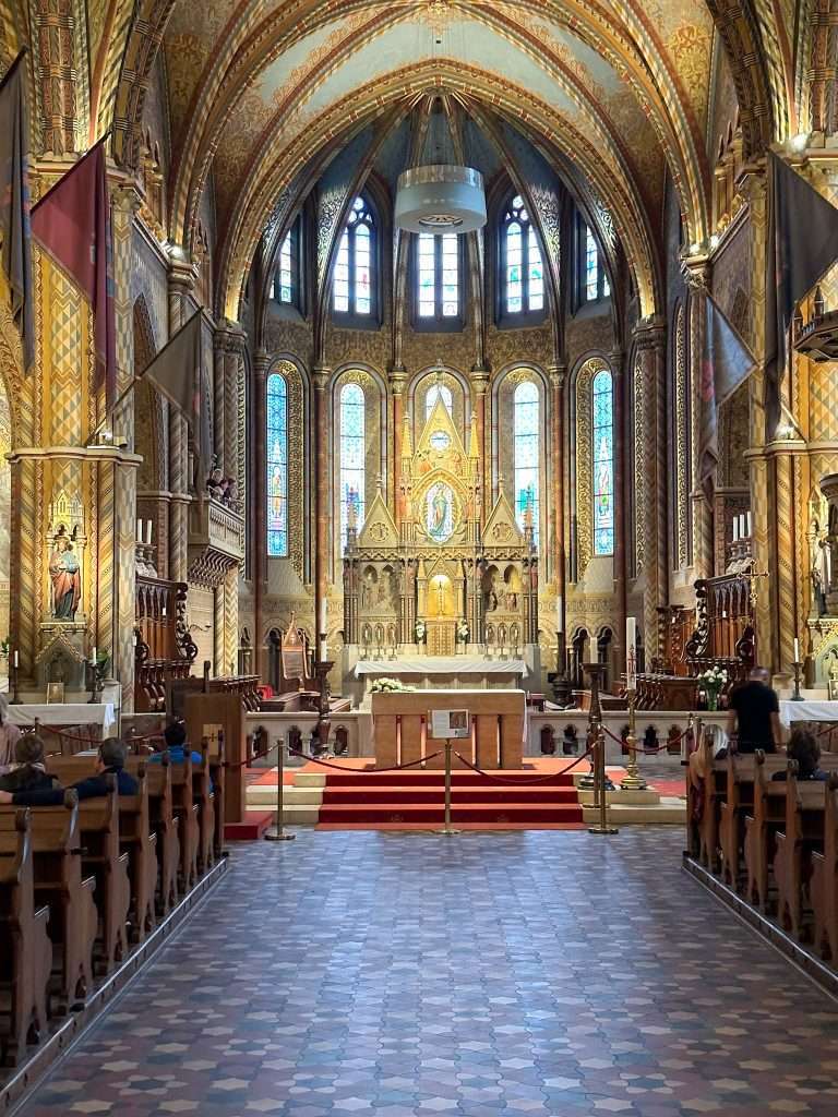 An interior view of part of Mátyás Church with typically gilded surfaces and wooden pews visible. 
