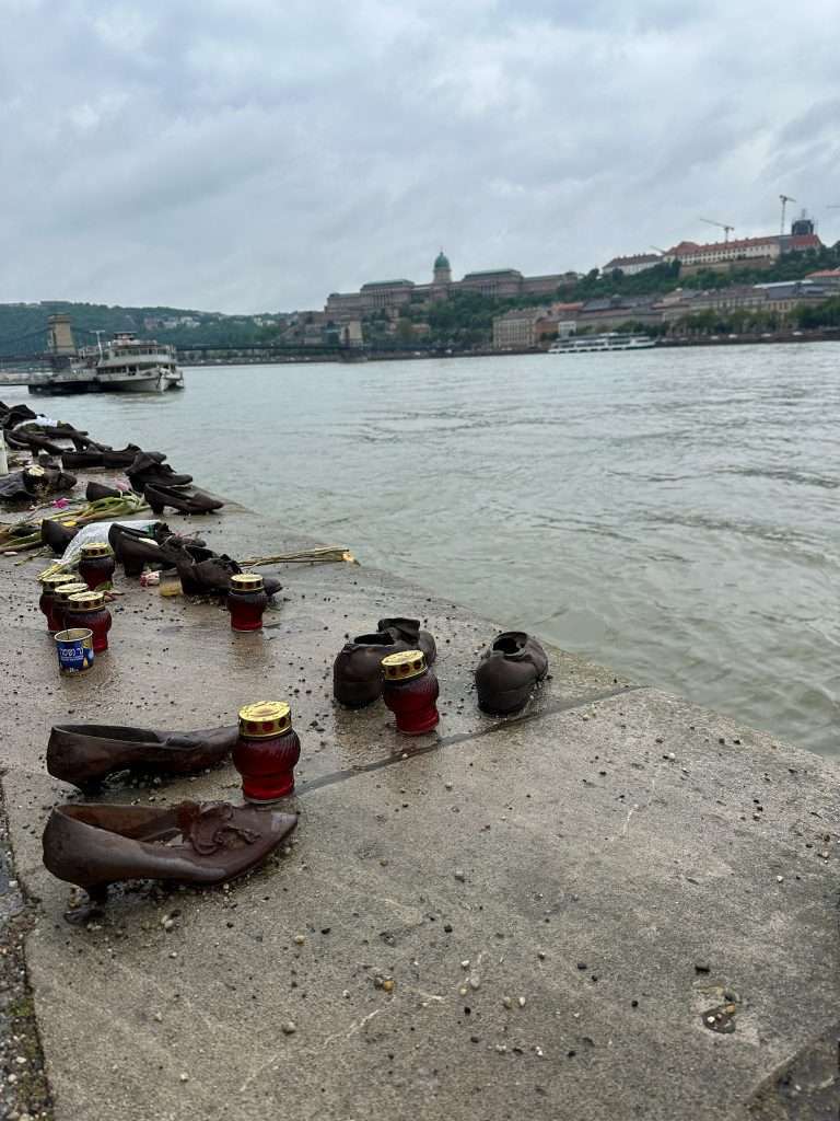 Cast iron shoes honouring Jews murdered in Budapest in 1944-45 are located along the banks of the River Danube. Amongst the shoes are candles, flowers and small offerings left by visitors to the Shoes on the Danube Memorial. The waters of the River Danube are visible in the image as is a boat in the background. 