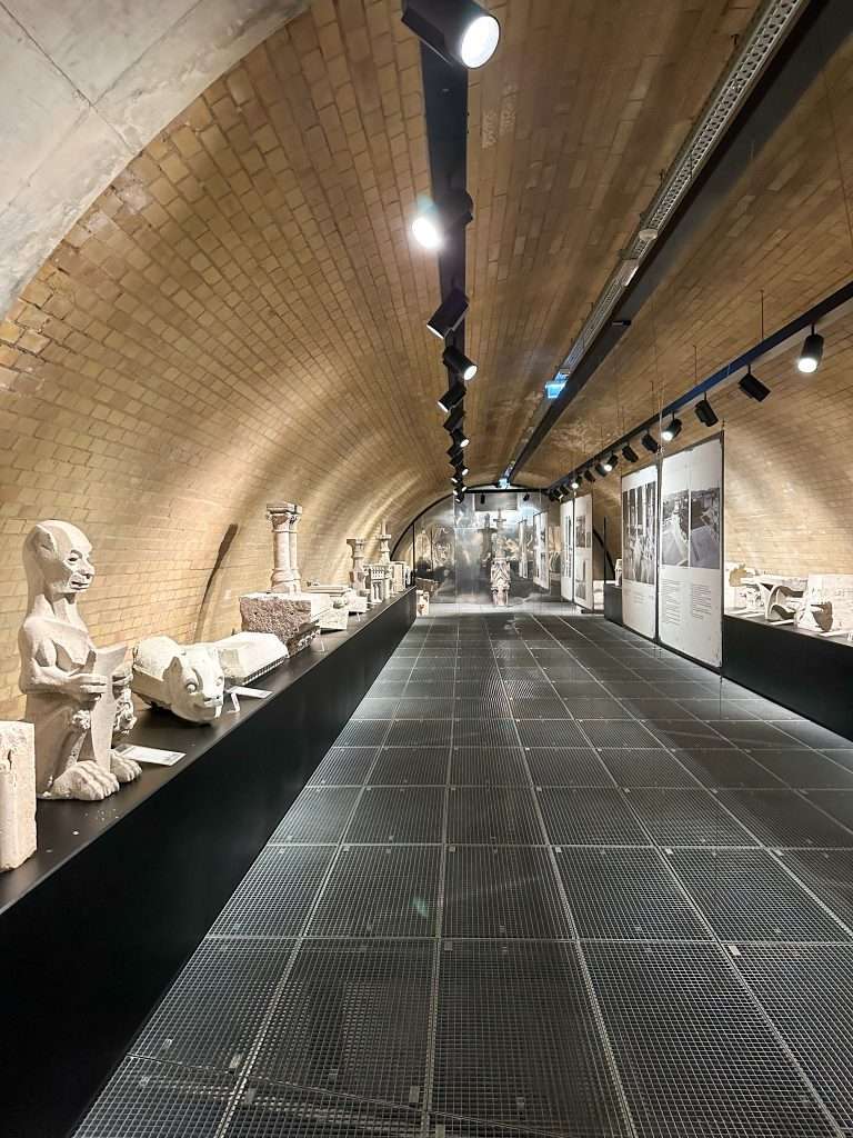 A view inside the Lapidarium museum in Budapest. Various characters carved from stone are displayed on long black plinths either side of a central, black coloured walkway. 
