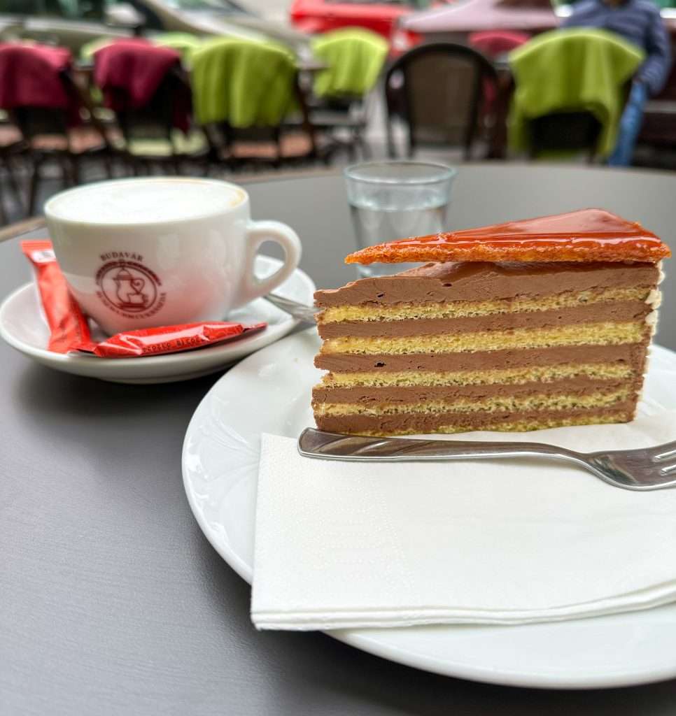 A slice of Dobos cake, a layered sponge cake with chocolate buttercream and a caramelised brown sugar topping, rests on a white plate on a cafe table at Ruszwurm confectionery in Budapest. A white coffee cup sits alongside it. A glass of water is visible in the background. 