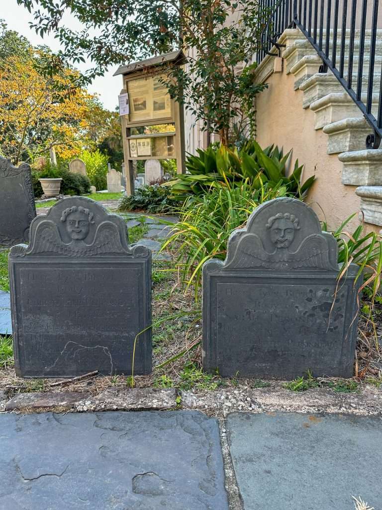 A view during day light hours of old headstones in a graveyard in Charleston South Carolina. Angel wings and faces are visible on the headstones. In the background are various trees and shrubs within the graveyard along with more headstones of different sizes and shapes. 