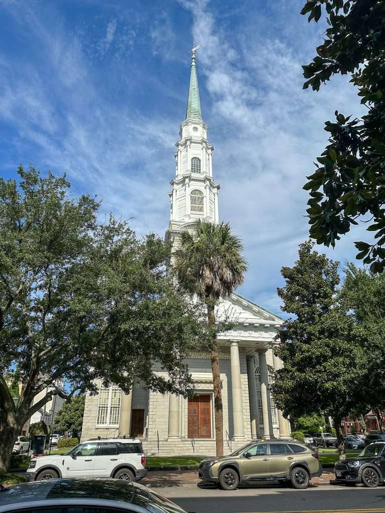 The Independent Presbyterian church with its pale cream exterior and white steeple sits under a blue but cloudy Savannah sky. Mature trees are visible growing on the pavements outside the church and several cars are seen parked in the street to the front of the church. 