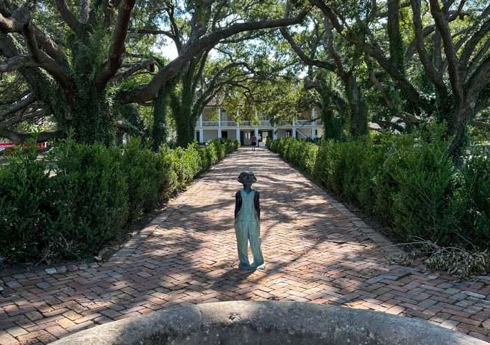 A view looking toward the large plantation house on the Whitney Plantation with the carved figure of a young enslaved child in the foreground.