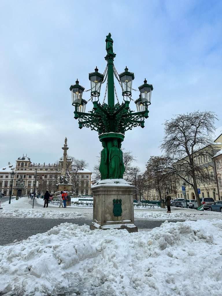 An impressive green iron lamp with 6 lanterns visible sits on a street outside Prague Castle. Piles of old fallen snow are visible in the image.
