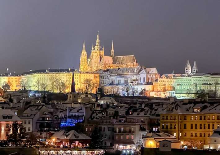 Prague Castle is lit up at night with various colours including gold, green and lavender visible. The image was captured from the Charles Bridge with the river visible in the foreground. The roofs of buildings on the river bank are dusted with snow.