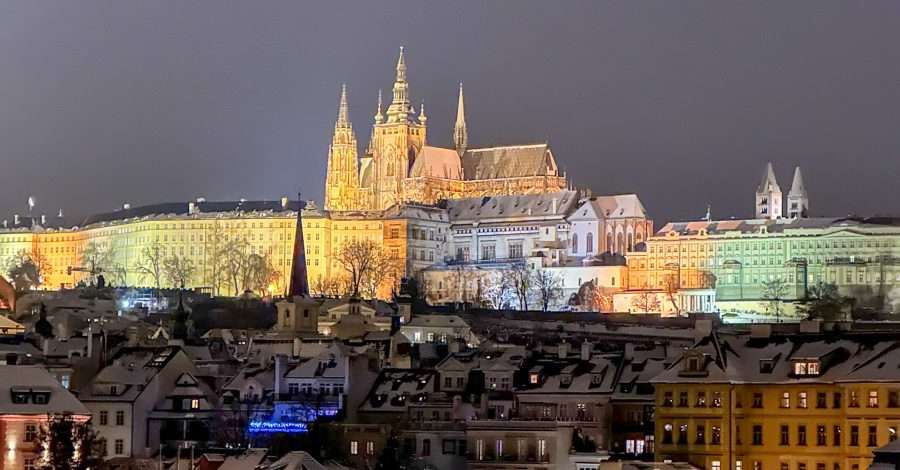 Prague Castle is lit up at night with various colours including gold, green and lavender visible. The image was captured from the Charles Bridge with the river visible in the foreground. The roofs of buildings on the river bank are dusted with snow.