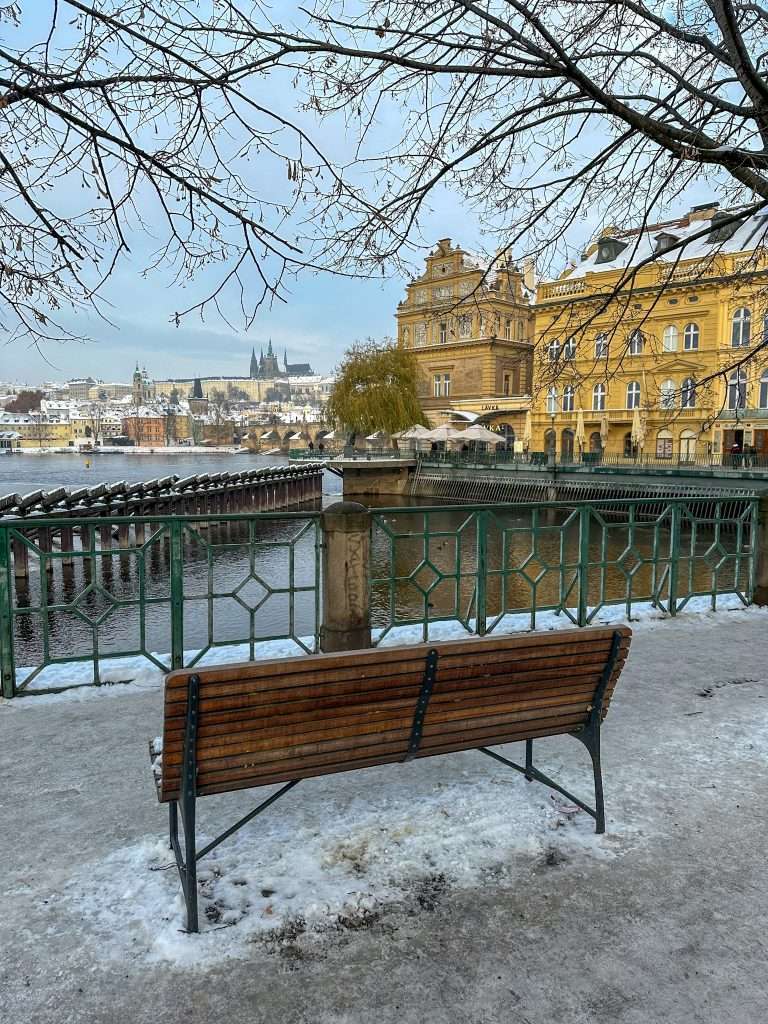 A view overlooking the Charles River toward Prague Castle. A wooden park bench is in the foreground with snow and ice visible on the ground.