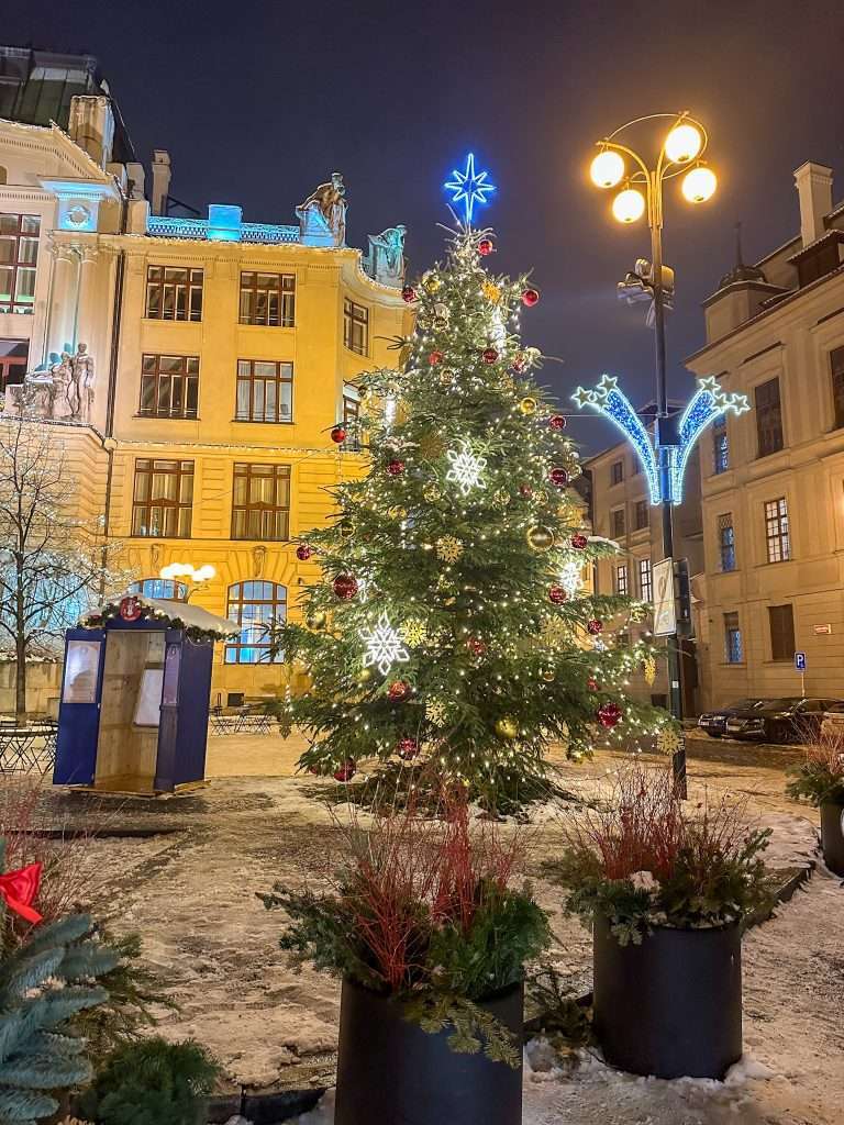 Image shows a scene at night in Prague during the time of the Prague Christmas Markets. The image shows a brightly lit Christmas tree surrounded by snow.