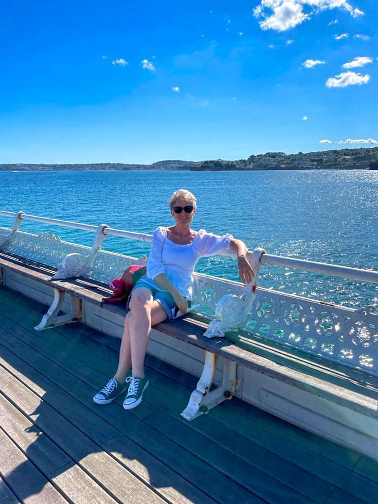 The author of this blog posts sits on a wooden bench against white coloured iron railings on Torquay Harbour. She is wearing sunglasses, a white short and shorts.