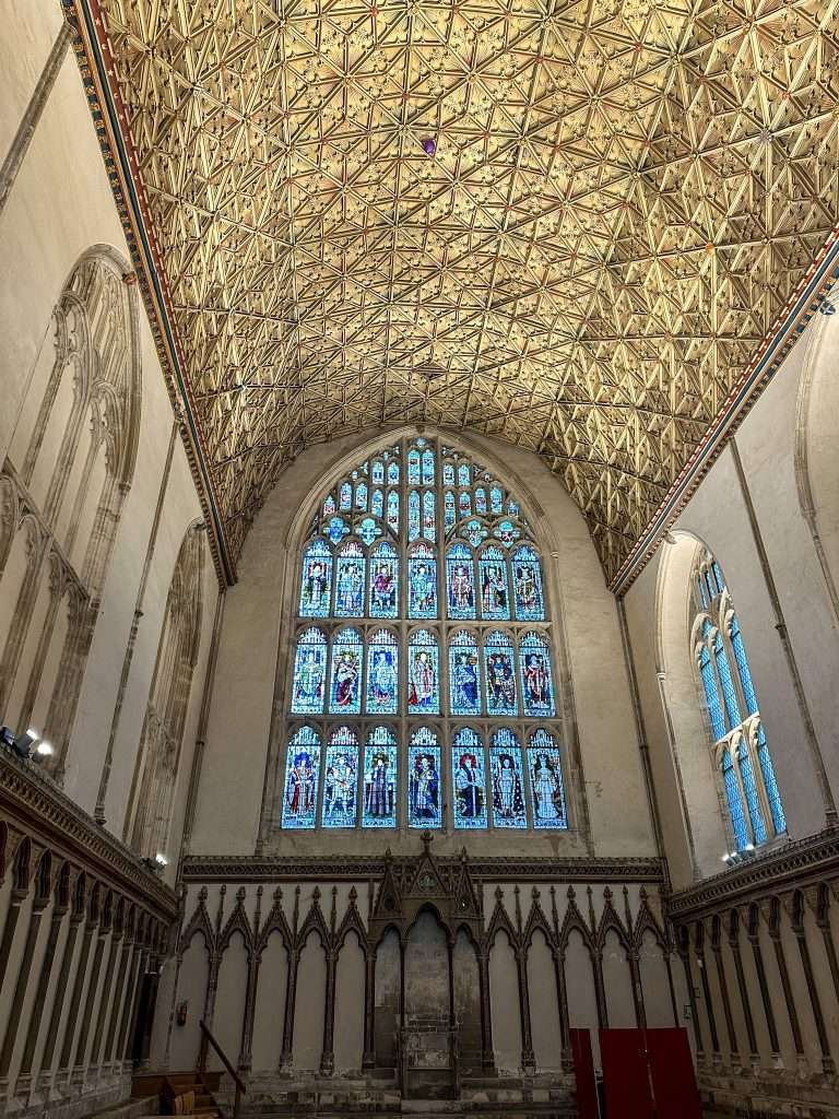 The Chapter House at Canterbury Cathedral with its highly decorated roof dating from 1400 and with one end dominated by stained glass windows  in an arch shape.