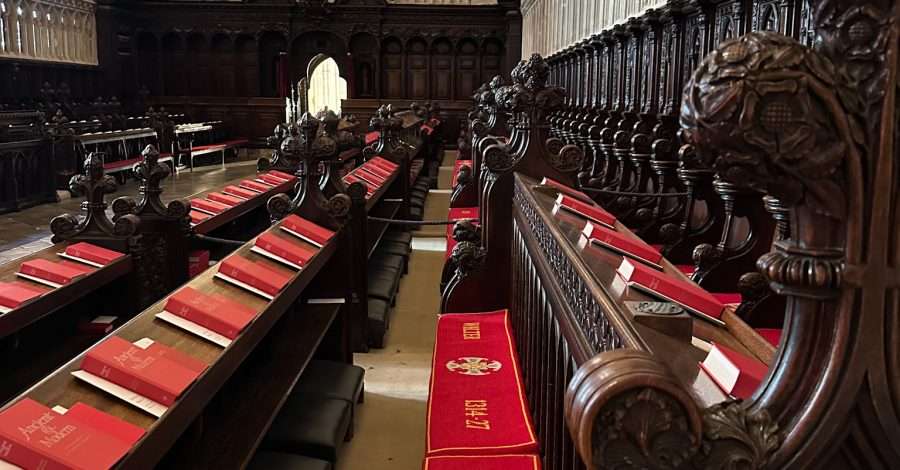 A view of some of the beautifully carved wooden pews inside the Quire within Canterbury Cathedral. Red bibles and cushions standout against the dark wood of the pews.