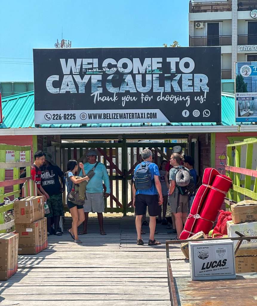 A "Welcome to Caye Caulker" sign in black and white greets arrivals to the island. A boardwalk is visible in the image as are several visitors. Various cardboard boxes are piled on the boardwalk, presumably awaiting their owners.