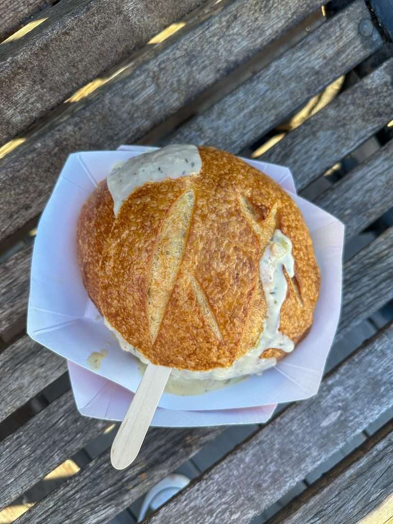 A serving of clam chowder sits within a sourdough bread bowl. The top of the bread bowl is in place but chowder can be seen running down the side of the bread bowl. The handle of a disposable wooden spoon is also visible. The sourdough bun and clam chowder are sitting in a cardboard box.