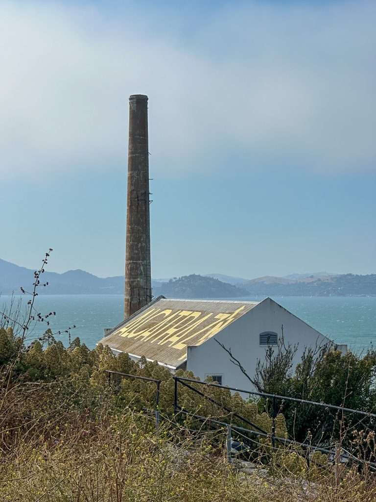A view of the powerhouse building on Alcatraz Island with a tall concrete chimney visible and the word Alcatraz written on yellow on the roof of the building. 