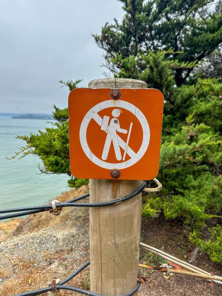 Image is of an orange and white coloured sign on the Lands End Trail. A hiker is pictured with a circle and line through the middle indicating that hikers should not go beyond this point.
